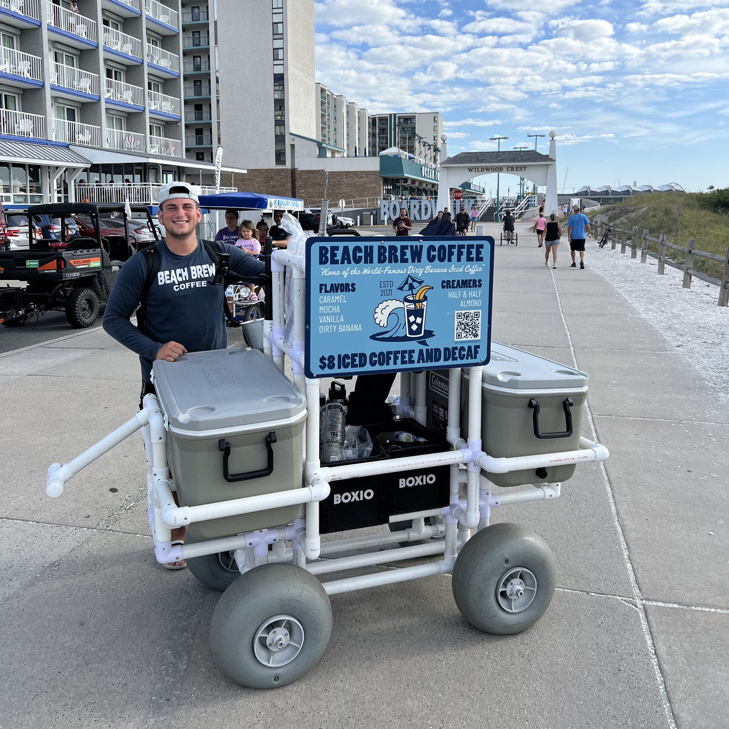 Person standing next to a 'Beach Brew Coffee' cart on a boardwalk with buildings and people in the background.